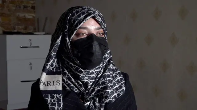 امرأة ترتدي حجاباً بلون أسود وأبيض وكمامة سوداء تجلس أمام الكاميرا