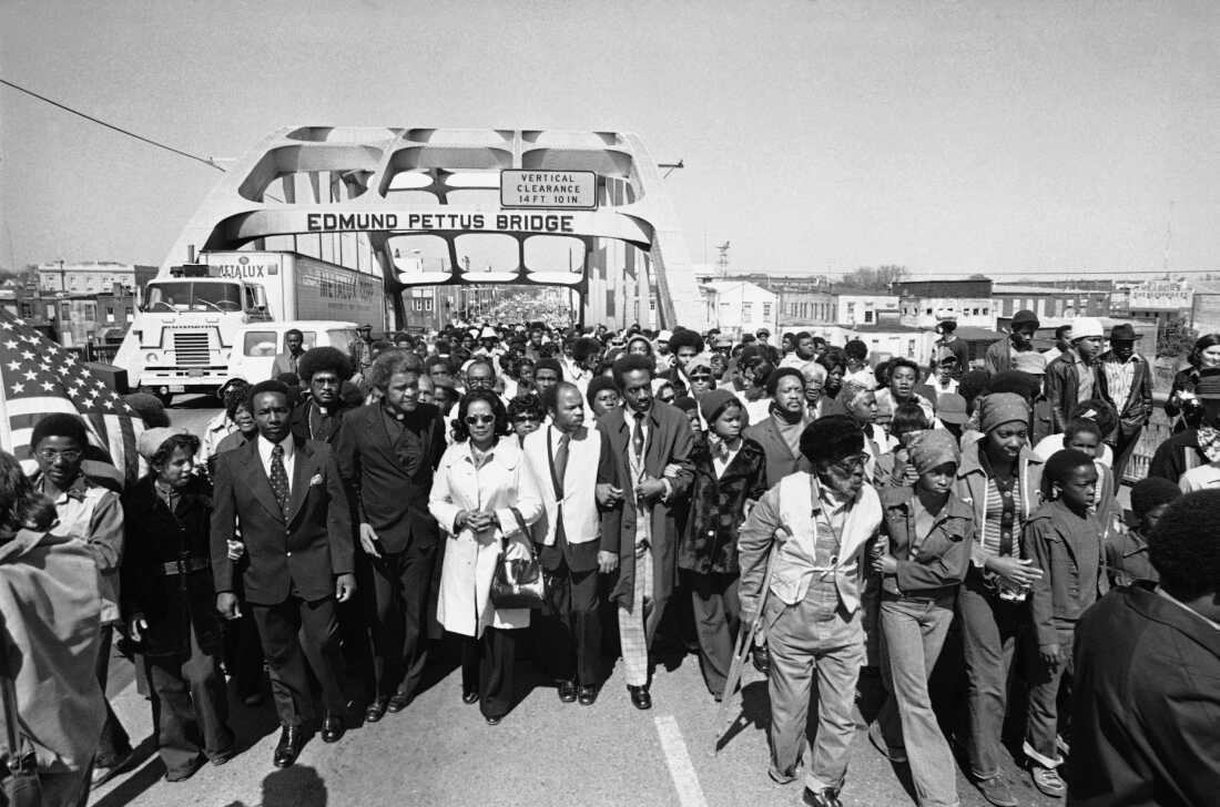 Aided by Father James Robinson, Mrs. Coretta Scott King, widow of Dr. Martin Luther King, Jr., center, and John Lewis of the Voter Education Project, a crowd estimated by police at 5,000, march across the Edmund Pettus Bridge from Selma, Alabama March 8, 1975.