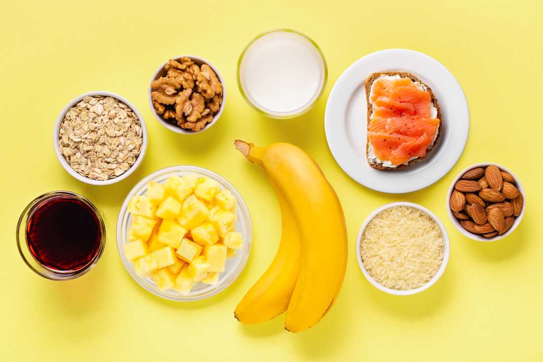 A high angle studio photograph of oats, walnuts, yogurt, lox on toast, almonds, brown rice, bananas, pineapple and cherry juice arranged on a yellow backdrop. 