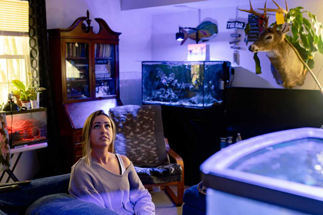 Becca Morris sits surrounded by fish tanks and plants, in the public housing apartment where she’s lived with her son for nine years, in Bridgeville, Delaware on Wednesday, May 14, 2025. (Wesley Lapointe/ for NPR)