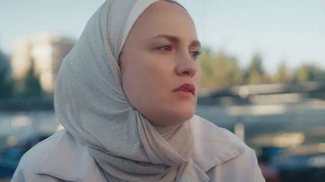 ترتدي ريم حجاباً رمادياً ناعماً ومعطفًا خفيفًا، وتنظر إلى مسافة متوسطة. 