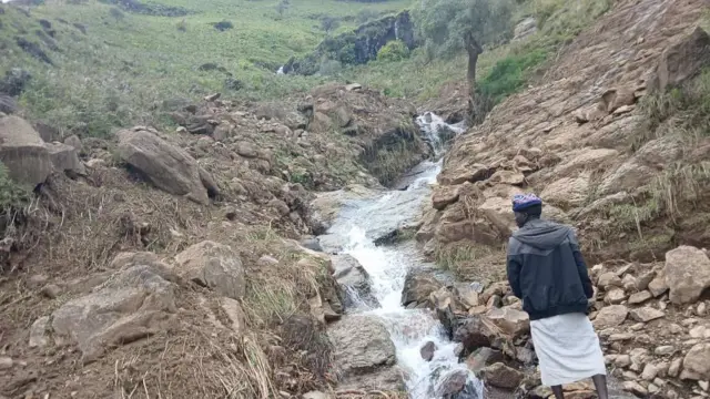 رجل يقف على حافة مجرى صخري في قرية ترسين بمنطقة جبل مَرَّة، ويرتدي جلابية بيضاء و سترة تجمع ما بين اللونين الأسود والرمادي