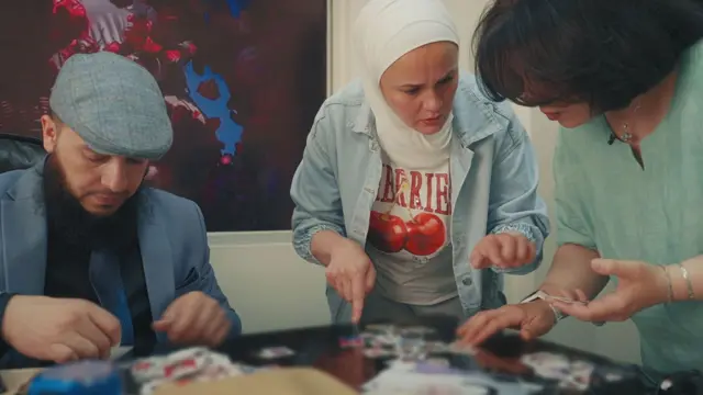 ترتدي مديرة دار رعاية الأطفال سترة زرقاء وقبعة تويد زرقاء. ريم ترتدي قميصاً أزرق مفتوحاً فوق تيشيرت عليه كرز، وشعرها مغطى بحجاب أبيض. جميعهن يتصفحن صور الأطفال المتناثرة على مكتب. 