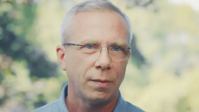 بينوا يرتدي نظارات بدون إطار، وعيون زرقاء، وشعرًا رمادياً، وقميصاً أزرق. 