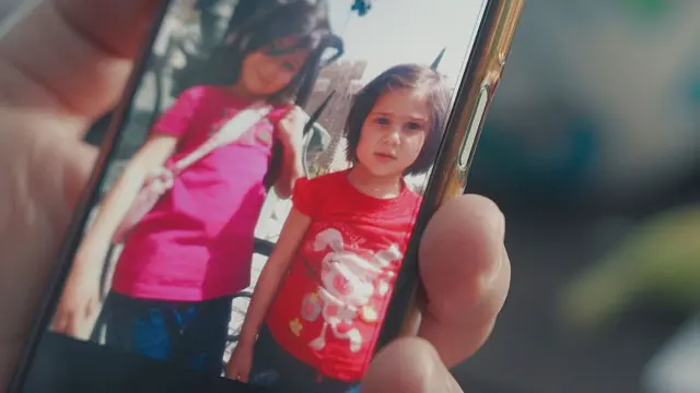 تبتسم طفلتان صغيرتان بشعر داكن وقميصين ورديين وأحمرين للكاميرا في صورة عُرضت على هاتف محمول. 