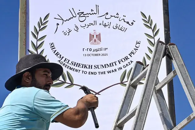 عامل بلدية يُصلح لافتة في شرم الشيخ، يرتدي قبعة سوداء وقميصاً بأكمام قصيرة لونه أزرق، يحمل مطرقة ويقف على سلم. كتب على اللافتة باللغتين العربية والإنجليزية: "قمة شرم الشيخ للسلام. اتفاق إنهاء الحرب في غزة". 