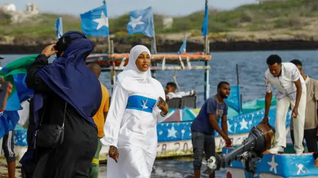 امرأة ترتدي حجاباً أبيض وفستاناً يمزج بين اللونين الأبيض والأزرق من أعلام الصومال، بينما تسير بجوار القوارب الراسية على شاطئ البحر.
