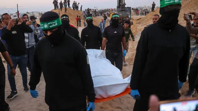 سكان من قطاع غزة يشاهدون مسلحين من كتائب عز الدين القسام، الجناح العسكري لحركة حماس وهم يحملون جثةً رهينة إسرائيلي انتشلت من نفقٍ في منطقة بشمال خان يونس جنوب قطاع غزة، في 28 أكتوبر/تشرين الأول 2025.