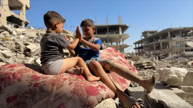 صورة تُظهر صبيين صغيرين، أحدهما بساق اصطناعية، يصفقان بأيديهما معاً أثناء جلوسهما على سجادة فوق أنقاض مبنى مدمر في غزة.
