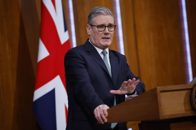 رئيس الوزراء البريطاني كير ستارمر متحدثاً، ومرتدياً بدلة رسمية كحلية اللون وربطة عنق تميل إلى اللون الرمادي ويبدو أنها منقطة باللون الأزرق، مع قميص أبيض. وخلفه العلم البريطاني.