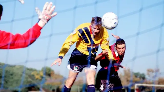 لاعب كرة قدم يسدد كرة برأسه وسط متابعة من لاعب من فريق منافس ويظهر أن الحارس يسعى للتصدي لها